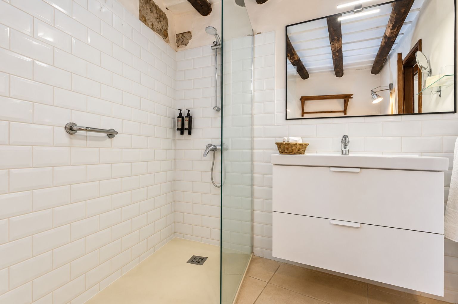 A bathroom with white tiled walls, a glass shower enclosure, a white vanity with a mirror, and a wooden beam ceiling.