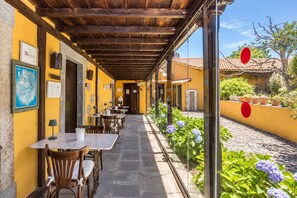 Ein Straßencafé mit Tischen und Stühlen, einer Holzpergola und Blick auf ein gelbes Gebäude mit Ziegeldach.