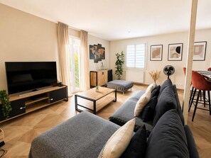 A modern living room with a flat-screen TV, grey sofas, a coffee table, and a dining area with a red chair. - Villa Bartholdi – House & Garden in Holtzheim (Holtzheim)