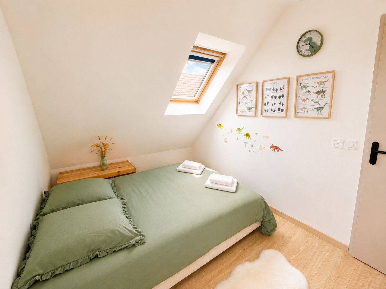 Ein Schlafzimmer mit Oberlichter, einem Bett mit grüner Bettwäsche, einem kleinen Nachttisch mit einer Vase und Wandstickern zum Thema Dinosaurier.