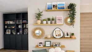 A room with a bookshelf, wall-mounted shelves with decorative items, a mirror, and a large flower-shaped cushion. - Villa Bartholdi – House & Garden in Holtzheim (Holtzheim)
