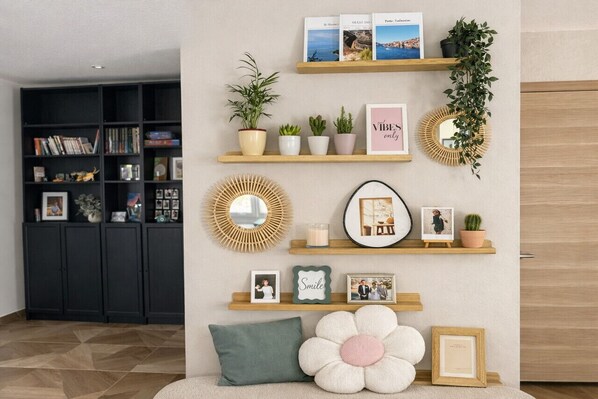 A room with a bookshelf, wall-mounted shelves with decorative items, a mirror, and a large flower-shaped cushion. - Villa Bartholdi – House & Garden in Holtzheim (Holtzheim)