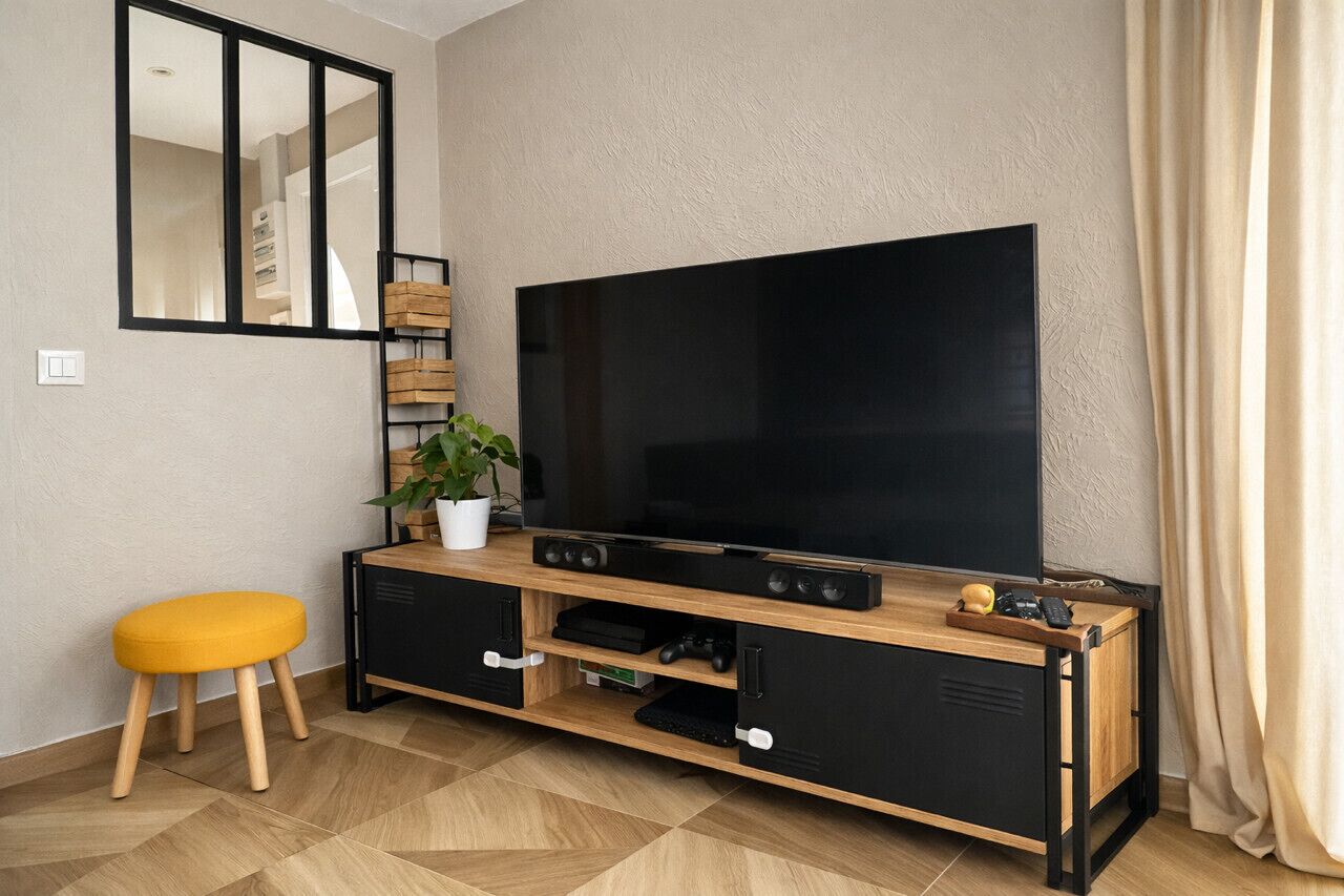 A living room with a large flat-screen TV, wooden entertainment unit, a yellow stool, and a potted plant.