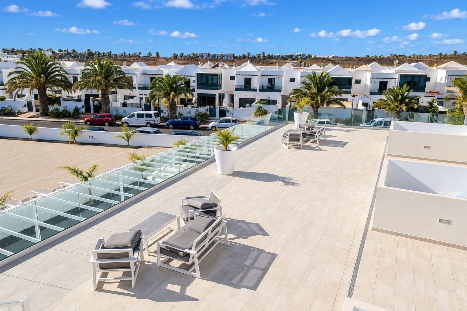 A rooftop terrace with outdoor seating and a glass balustrade overlooking a parking area and a row of white buildings.