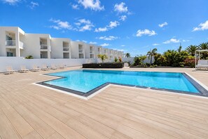 A swimming pool with a wooden deck, white lounge chairs, and a white building with balconies in the background. - GuestReady - Senator Terrace Retreat (Costa Teguise)