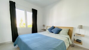 A bedroom with a bed, a window with curtains, a bedside table with a lamp, and a wall-mounted shelf. - GuestReady - Senator Terrace Retreat (Costa Teguise)