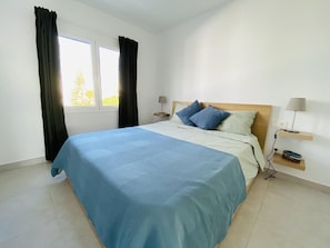 Ein Schlafzimmer mit Bett, Fenster mit Vorhängen, Nachttisch mit Lampe und einer Wandregal.