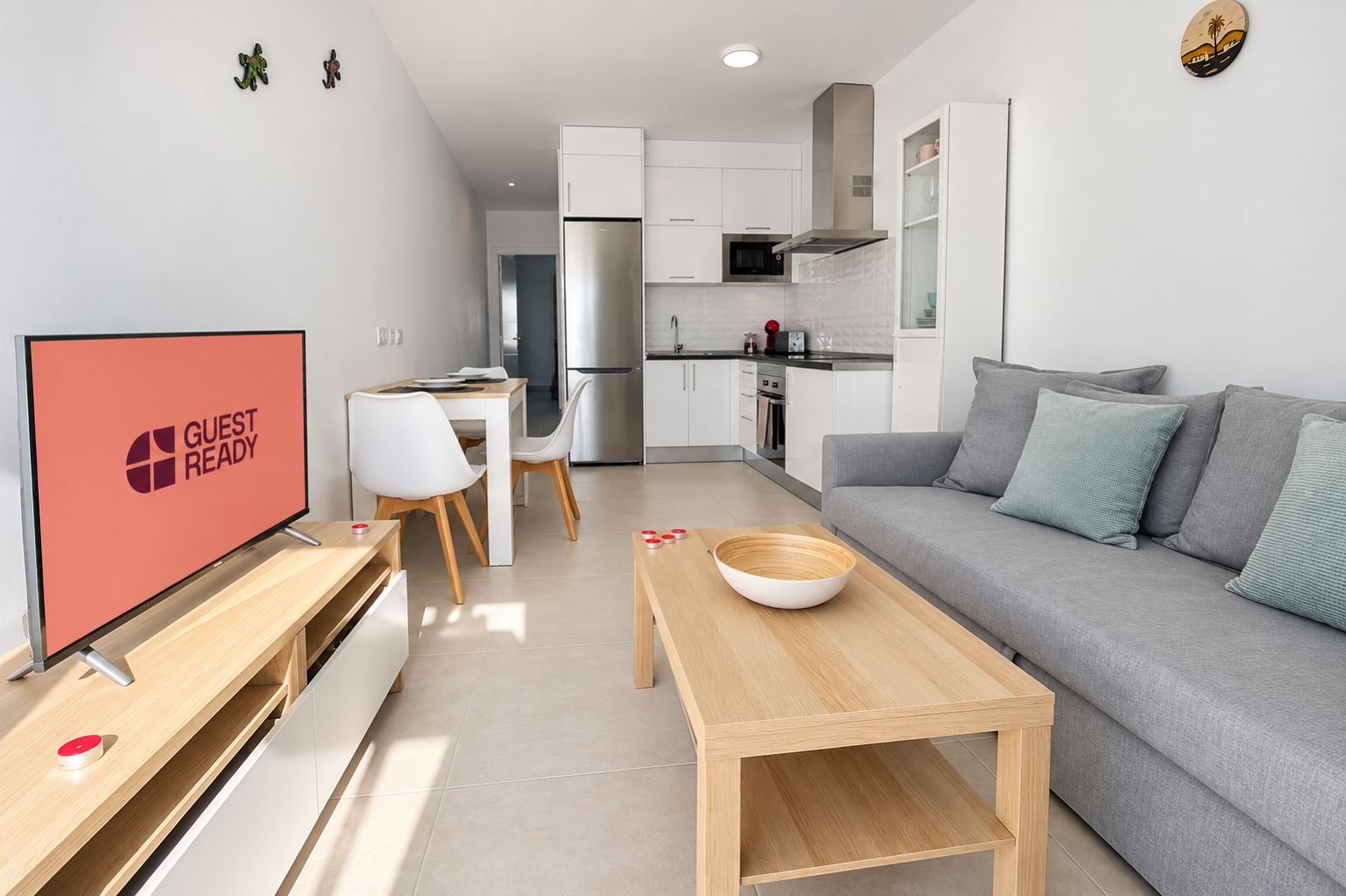 A modern living room with a flat-screen TV, a grey sofa, a wooden coffee table, and a dining area with a white table and chairs.