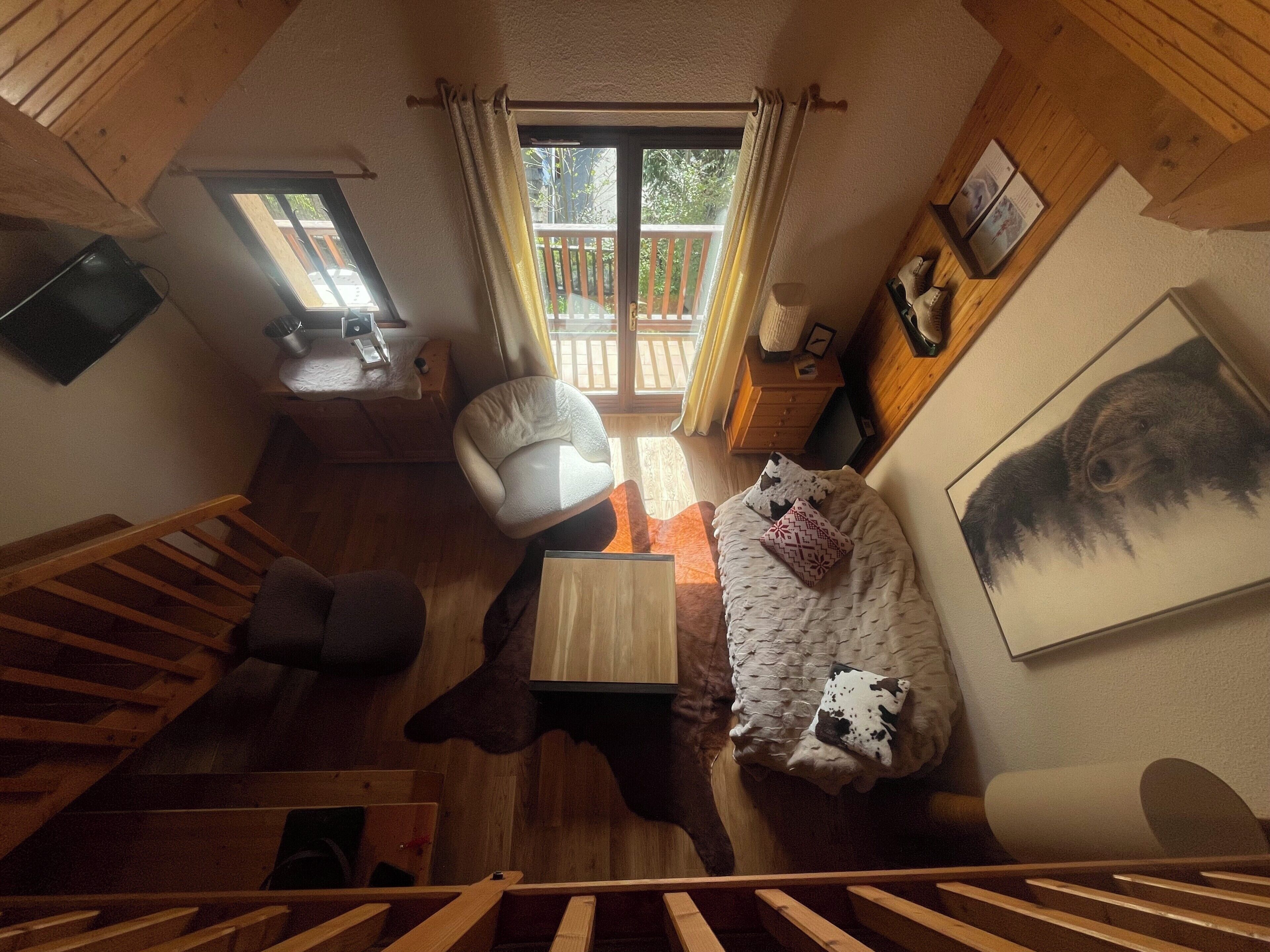 A loft-style room with a wooden staircase, a sofa, a chair, a small table, and a large window with curtains.