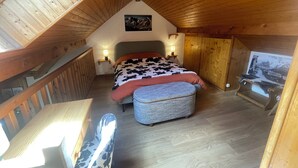 Une chambre mansardée chaleureuse avec un plafond en bois, un lit recouvert d’une couverture à motifs de vaches, une petite table et un banc.