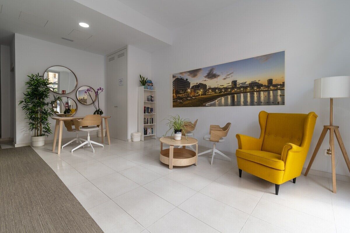 A modern living room with a yellow armchair, a round coffee table, a bookshelf, a mirror, and a cityscape painting on the wall.