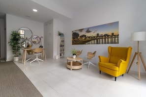 A modern living room with a yellow armchair, a round coffee table, a bookshelf, a mirror, and a cityscape painting on the wall. - GuestReady - 05B Livian Suites Ext 4 pax (Las Palmas de Gran Canaria)