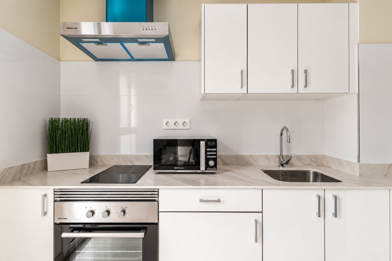 Eine moderne Küche mit weißen Schränken, einer Edelstahl-Dunstabzugshaube, einer Mikrowelle, einem Spülbecken mit modernem Wasserhahn und einem eingebauten Backofen.