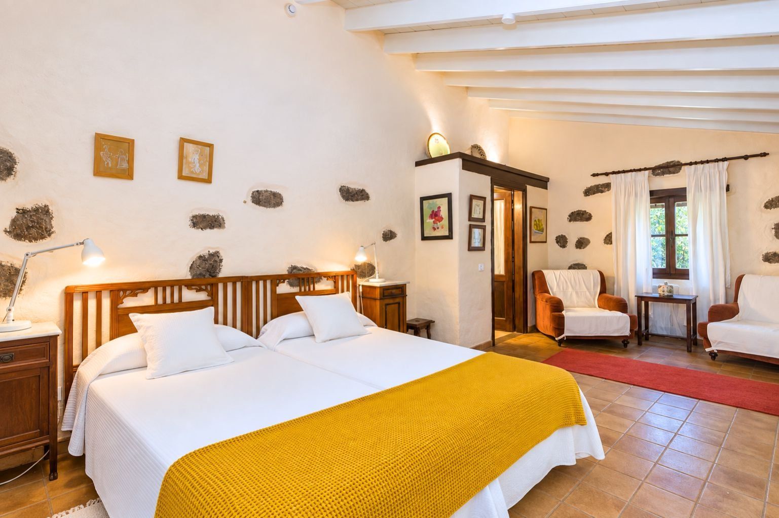 A hotel room with a double bed, a wooden headboard, a yellow throw blanket, a sitting area with two chairs, a small table, and a window with curtains.