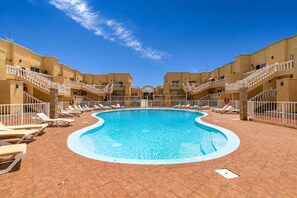 En swimmingpool med liggestole og en fliseterrasse, omgivet af høje bygninger med altaner.