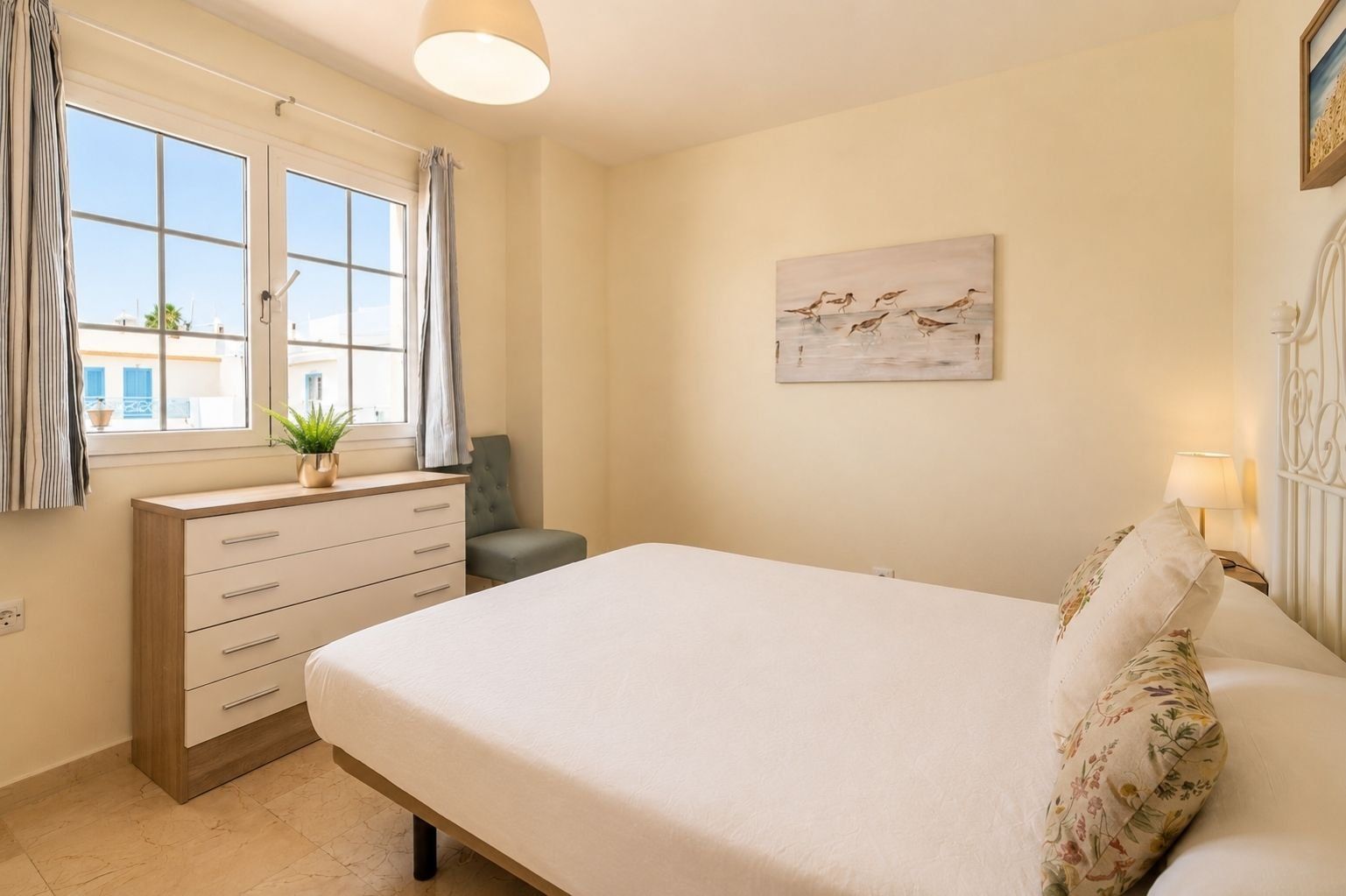 A bedroom with a bed, a dresser, a chair, a window with curtains, a painting on the wall, and a lamp on the nightstand.