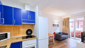 Een compacte keuken met blauwe kastjes, een witte koelkast, een magnetron en een fornuis. Er is een eethoek met een tafel en stoelen, en een bank.