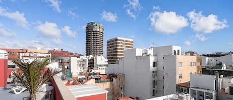 A rooftop terrace with a potted plant, a red wall, and a view of the cityscape including a tall building.