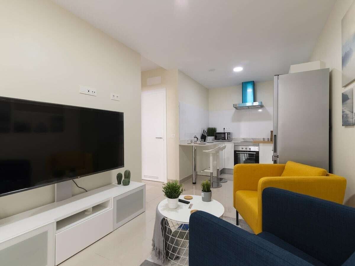 A living room with a flat-screen TV, a sofa, a coffee table, and a kitchen area in the background.
