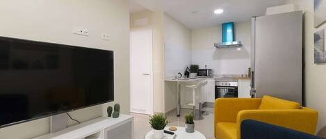 A living room with a flat-screen TV, a sofa, a coffee table, and a kitchen area in the background.