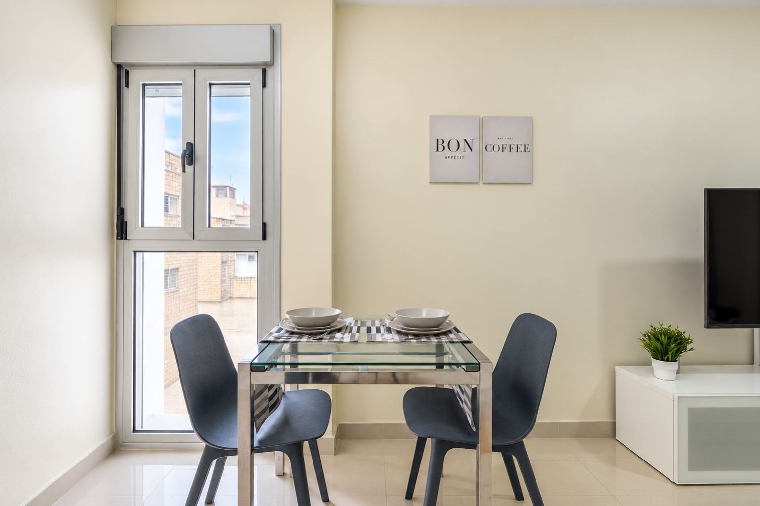 A modern dining area with a glass table, two chairs, and a television on a white cabinet.