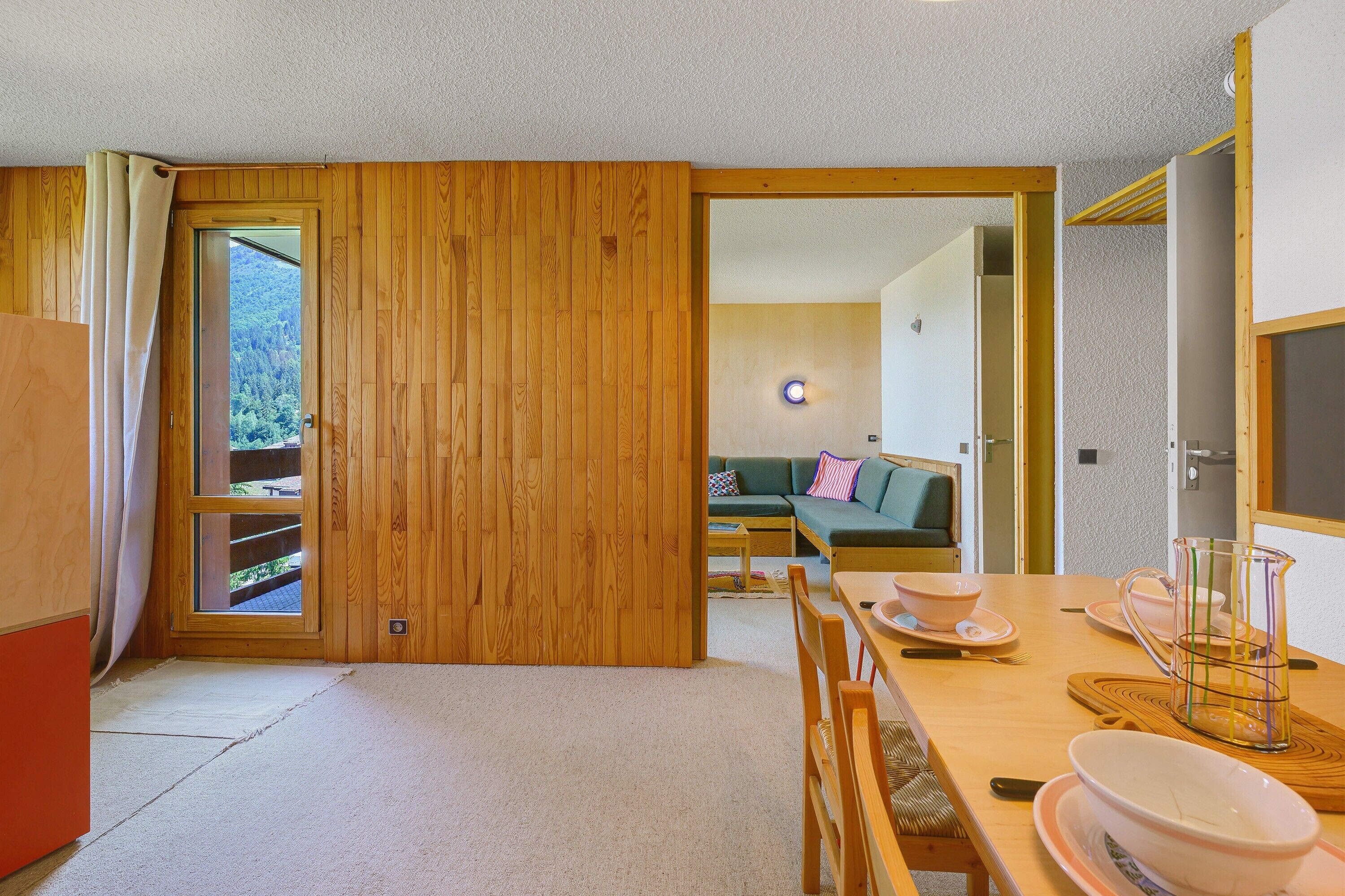 Ein Zimmer mit Holztür, einem Esstisch mit Stühlen, einer Couch und Blick auf einen Wald durch ein Fenster.