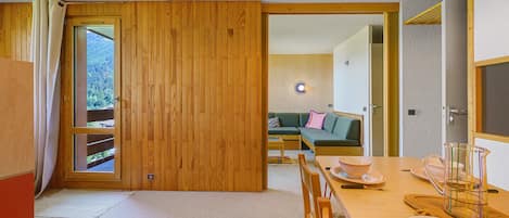 A room with a wooden door, a dining table with chairs, a sofa, and a view of a forest through a window.