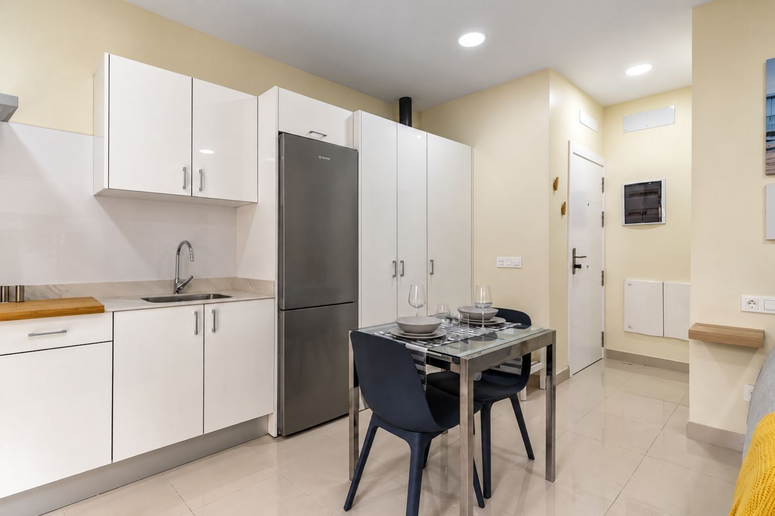 A modern kitchen with white cabinets, a stainless steel refrigerator, and a dining table set for two.