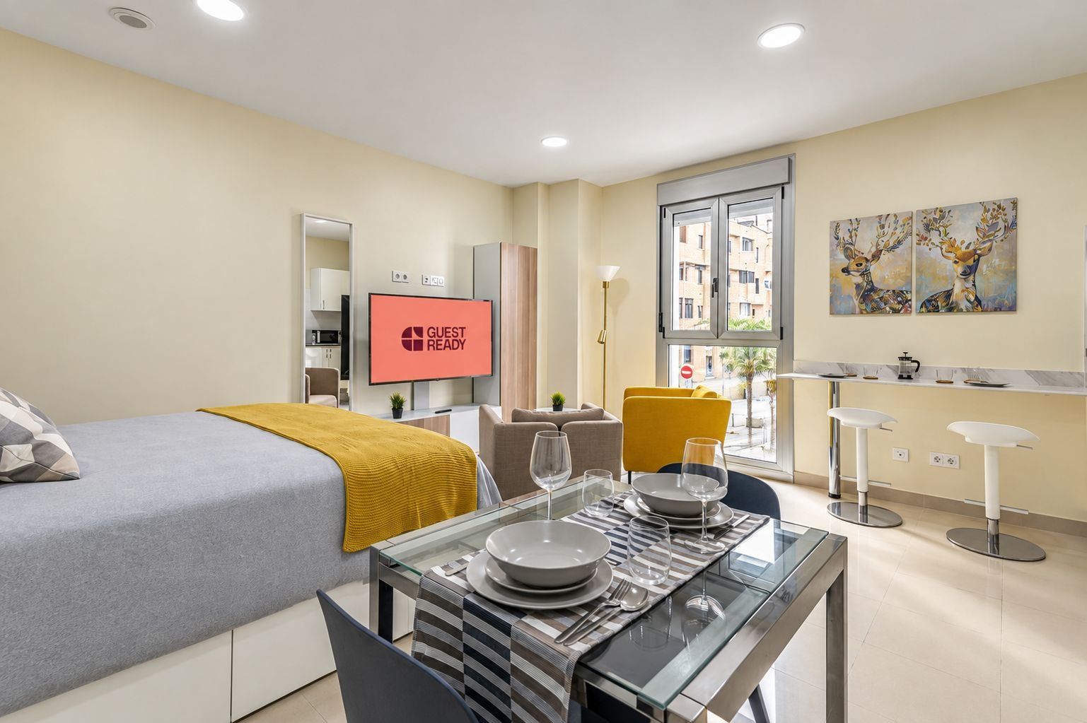 A hotel room with a bed, a TV displaying 'GUEST READY,' a dining area with a table set for two, and a window with a view of buildings.