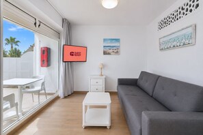 Ein modernes Wohnzimmer mit einem grauen Sofa, einem weißen Couchtisch, einem Fernseher, der 'GUEST READY' anzeigt, und einem Fenster mit Blick auf Palmen.