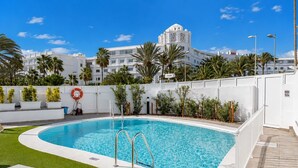 A swimming pool with a white deck, surrounded by palm trees and a white building with balconies in the background. - GuestReady - Alemania Villa Garden (San Bartolomé de Tirajana)