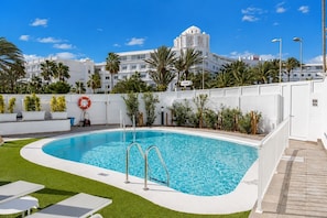 A swimming pool with a white deck, surrounded by palm trees and a white building with balconies in the background. - GuestReady - Alemania Villa Garden (San Bartolomé de Tirajana)