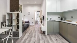 Una cocina moderna con armarios blancos, un comedor con mesa y sillas blancas, y una sala de estar con sofá y estantería.
