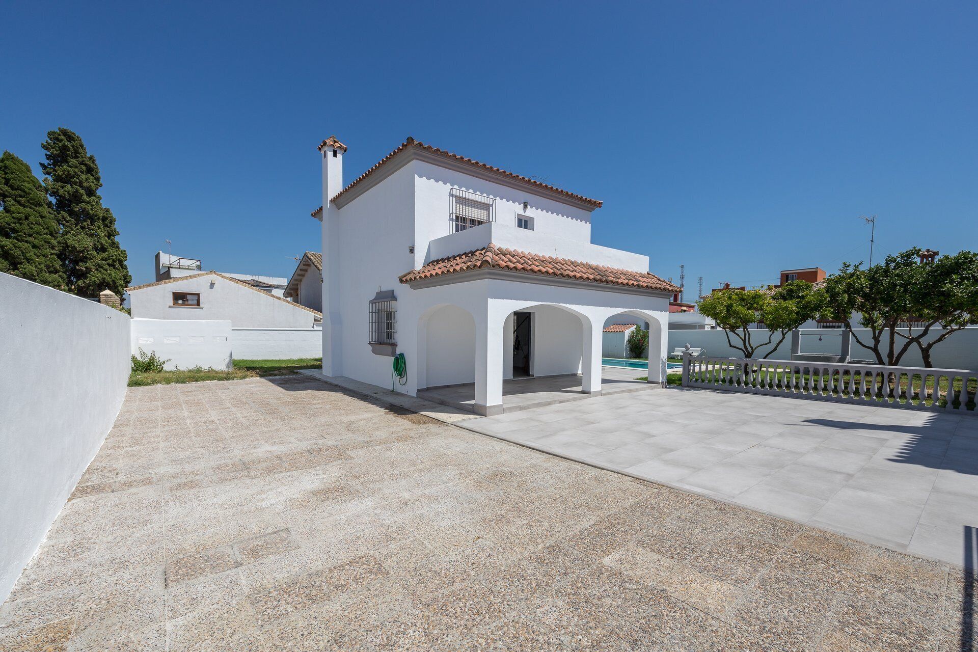 Un bâtiment blanc avec un toit recouvert de tuiles, un patio abrité et une piscine à l’arrière-plan.