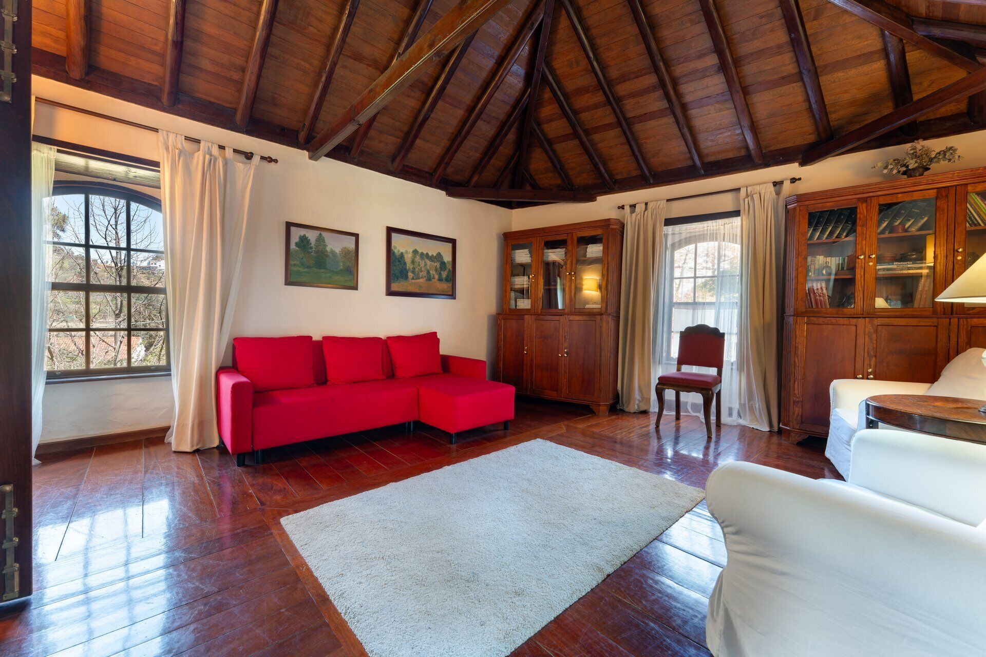A room with a wooden ceiling, a red sofa, a white armchair, a cabinet, and a window with curtains.
