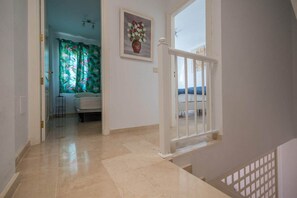 A hallway with a white railing, a door leading to a room with a bed, a painting on the wall, and a window with curtains. - GuestReady - Puerto Rico Family Retreat (Las Palmas de Gran Canaria)