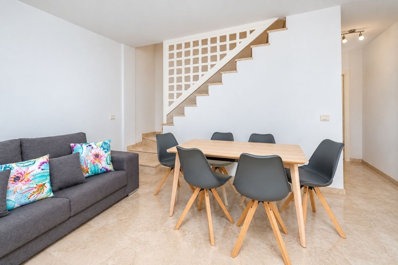 Una sala de estar moderna con un sofá gris, almohadones coloridos, una mesa de comedor de madera y una escalera con baranda de madera.