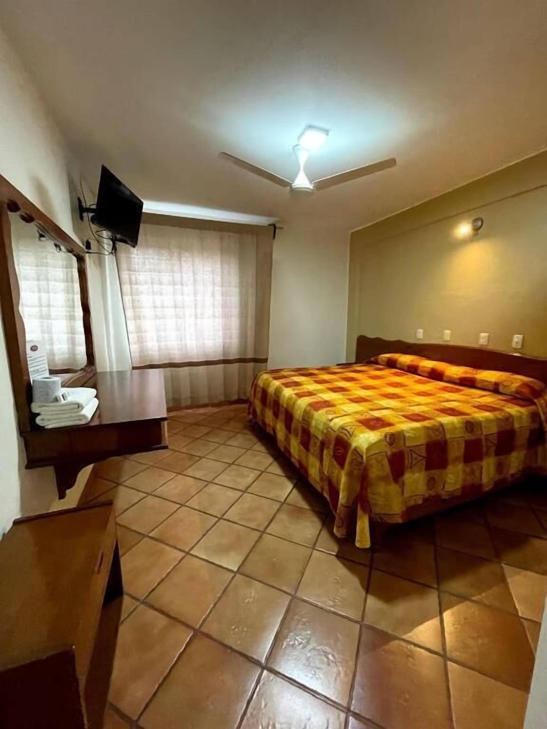Un dormitorio con una cama, ventilador de techo, televisor y ventana con cortinas.