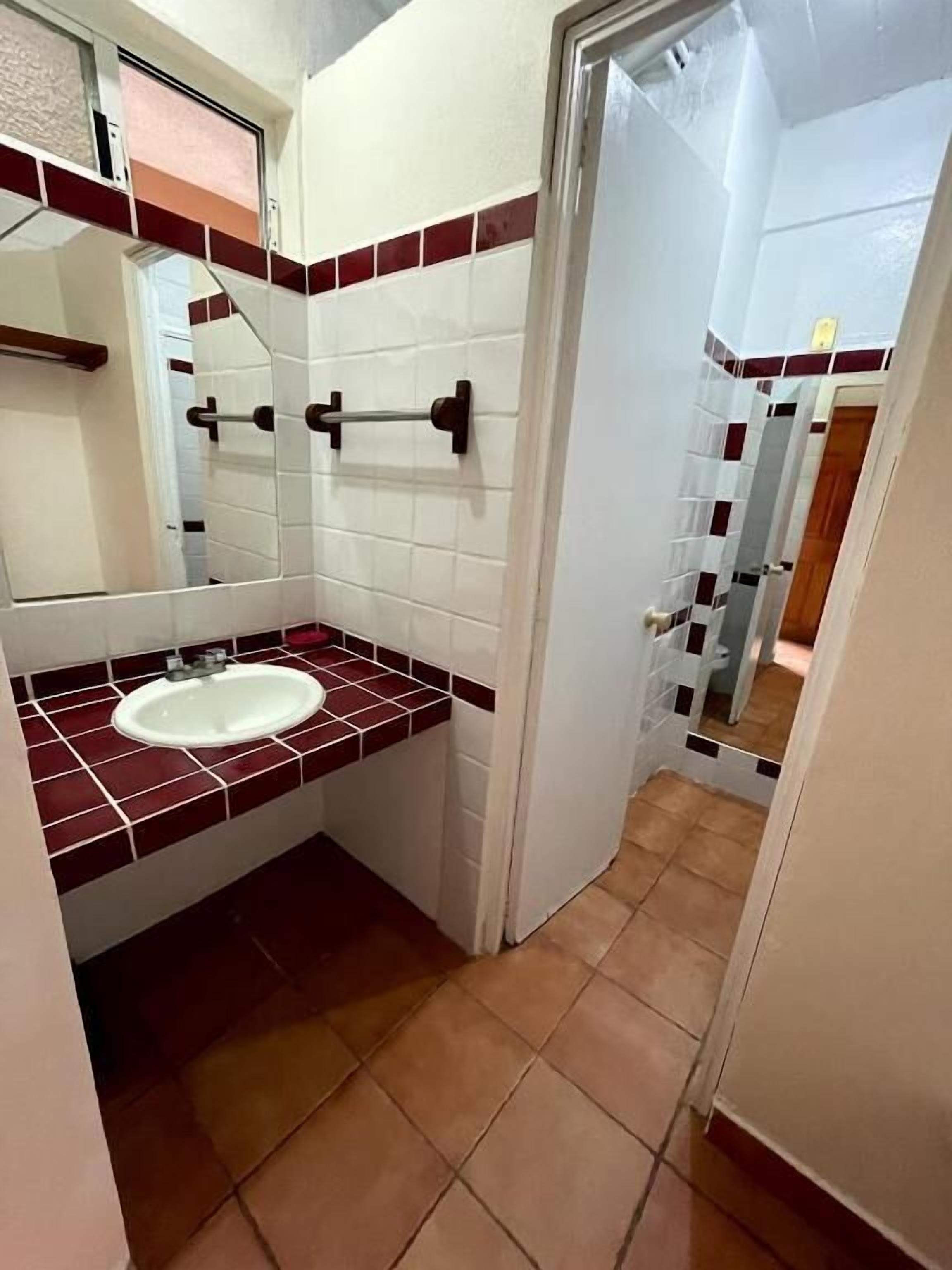 Un baño con un lavamanos blanco, baldosas rojas y blancas, un espejo y una puerta que da a otra habitación.