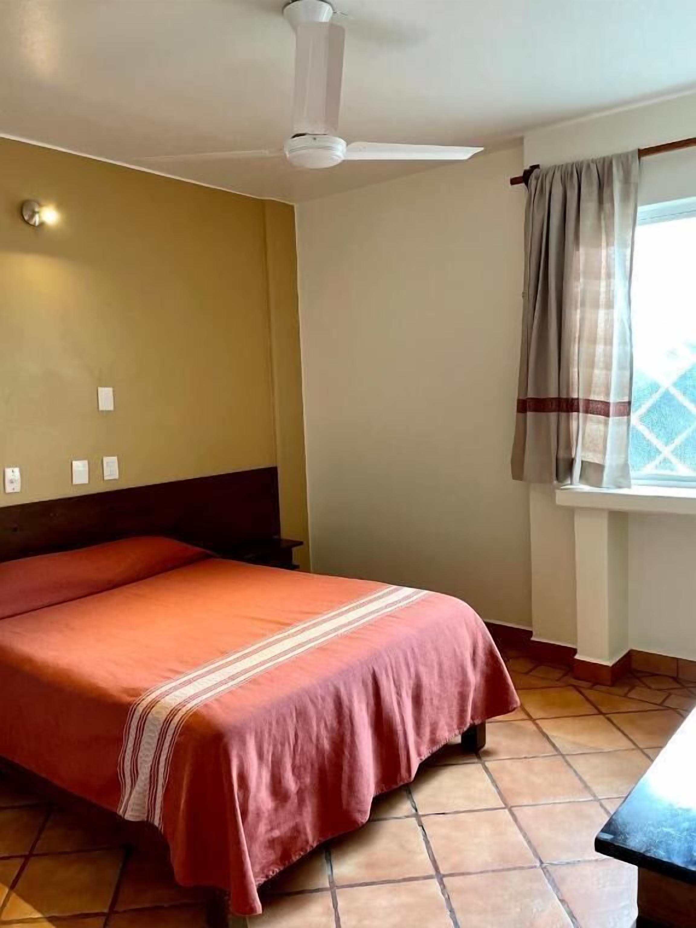 Una habitación con una cama, un ventilador de techo, una ventana con cortinas y suelo de baldosas.