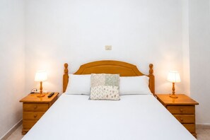 Ein ordentlich bezogenes Bett mit Holz-Kopfteil, zwei Nachttische mit Lampen und eine Steckdose an der Wand.