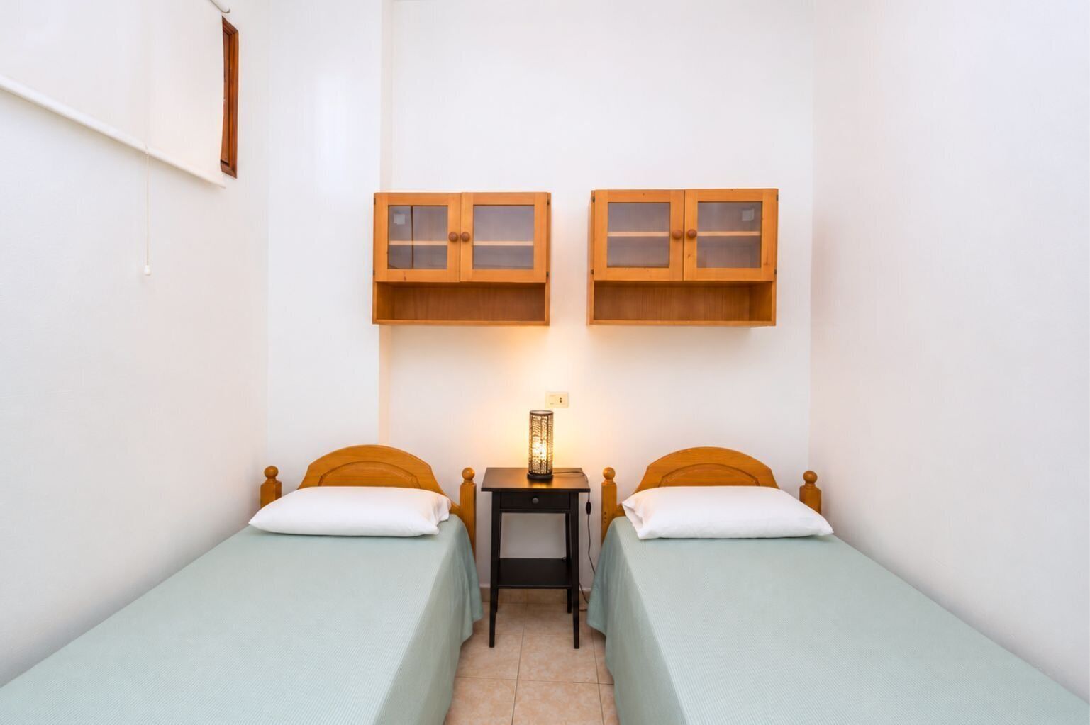 Une chambre avec deux lits simples, chacun doté d’une tête de lit en bois et d’une couvre-lit vert clair. Deux armoires en bois, avec des portes en verre, sont placées au-dessus des lits.