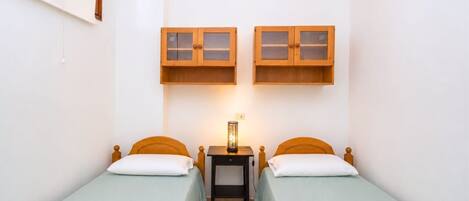 Ein Zimmer mit zwei Einzelbetten, jeweils mit einem Holzkopfstück und einer hellgrünen Bettdecke. Darüber befinden sich zwei Nachttische aus Holz mit Glastüren.