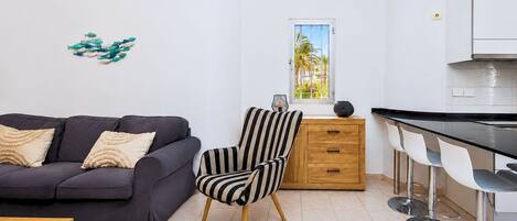 Una sala moderna con un sofá, un sillón a rayas, una mesita de madera y una zona de cocina con un taburete de bar.