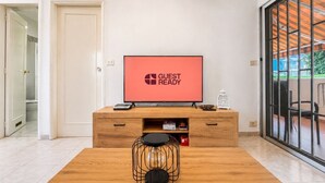 Una camera moderna con un mobile TV in legno, una TV a schermo piatto che mostra la scritta 'GUEST READY', un tavolino da caffè e una porta scorrevole in vetro che dà su una zona esterna.