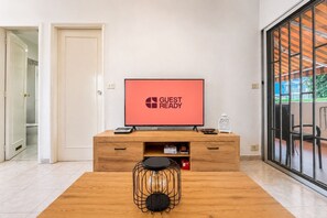 Ein modernes Zimmer mit einer Holz-TV-Konsole, einem Flachbildfernseher, der 'GUEST READY' anzeigt, einem Couchtisch und einer Glastür zum Außenbereich.