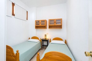Quarto pequeno e estreito com duas camas de madeira, uma mesa de cabeceira e dois armários de madeira sobre as camas.