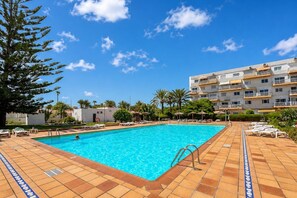 Ein rechteckiger Pool mit Fliesenbordur, umgeben von Liegestühlen und Sonnenschirmen, mit einem mehrstöckigen Gebäude im Hintergrund.