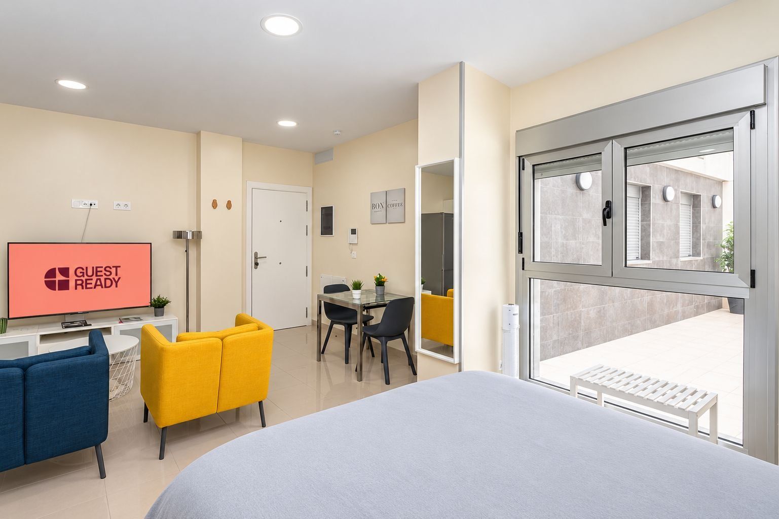 Una camera d'albergo con un letto, una scrivania con una sedia, una TV che mostra 'GUEST READY' e un balcone con vista.