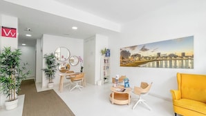 A modern hotel lobby with a seating area, a reception desk, and a large cityscape painting on the wall. - GuestReady - 01A Livian Suites 1A Int 2pax (Las Palmas de Gran Canaria)